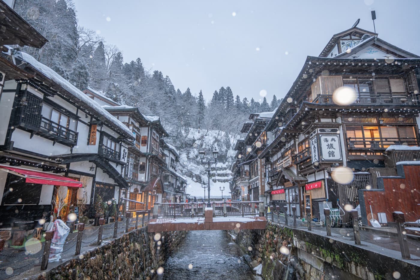 Ginzan Onsen's wooden ryokan street under falling snow in the morning, the river running between snow-covered banks