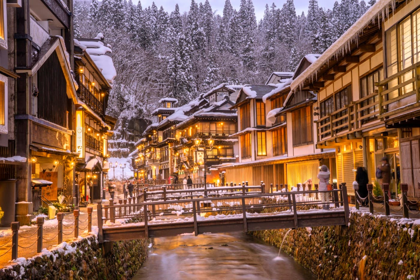 Ginzan Onsen at dusk in winter, gas lamps and ryokan windows glowing warm against deep snow along the river