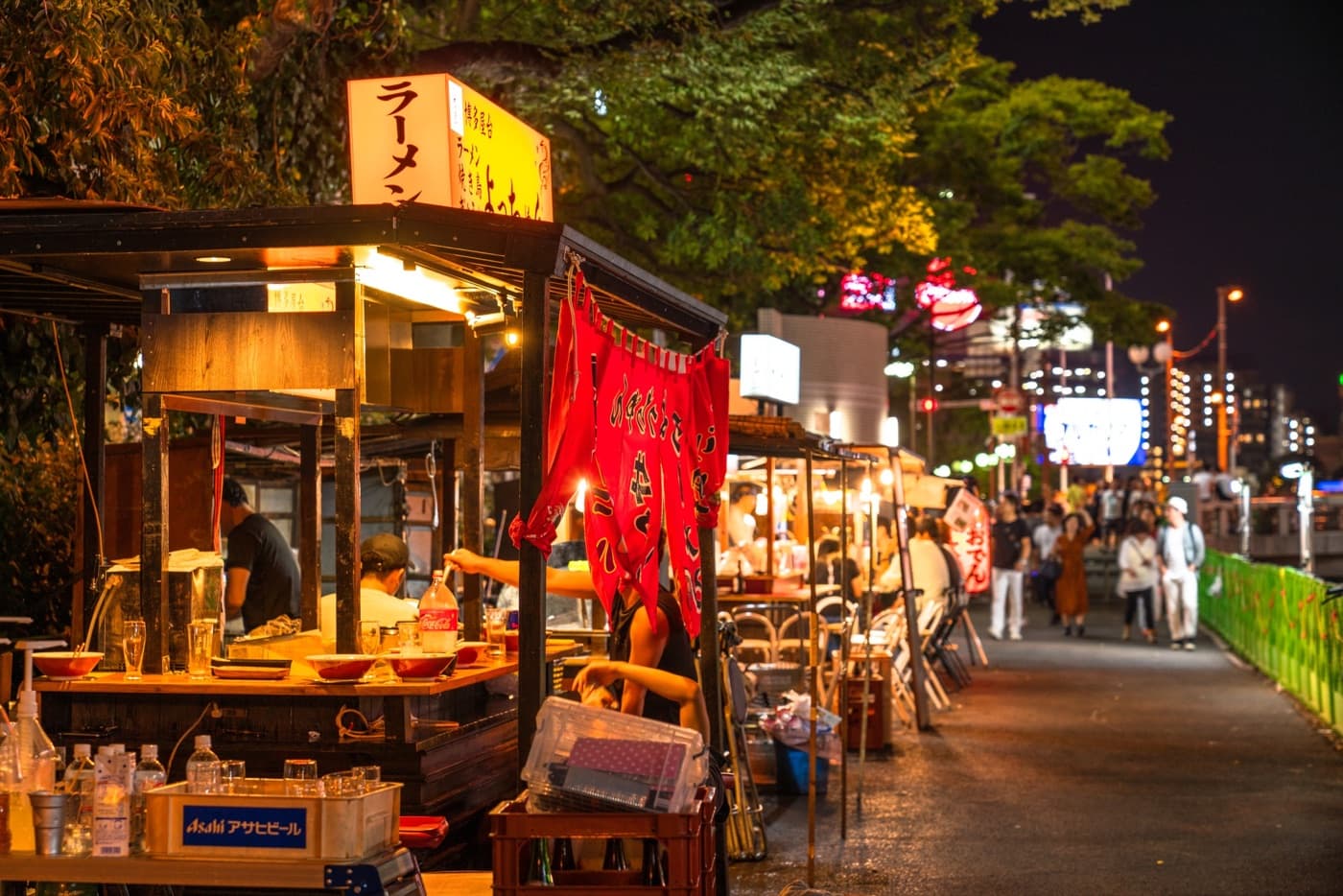 Yatai food stalls glowing at night along the river in Nakasu, Fukuoka, with red noren curtains and diners at the counters