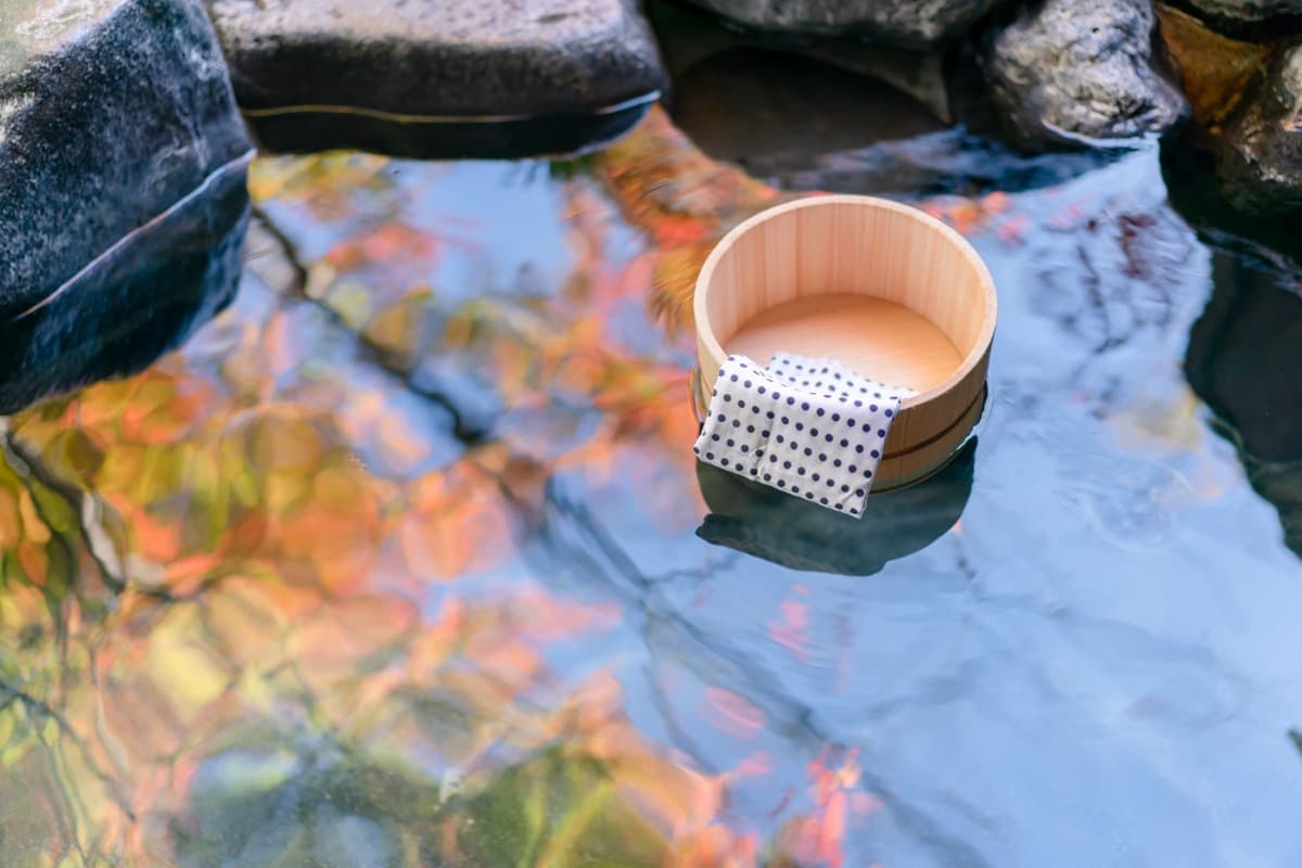 A wooden bath bucket and towel resting on the stones of an open-air hot spring, with autumn leaves reflected in the water