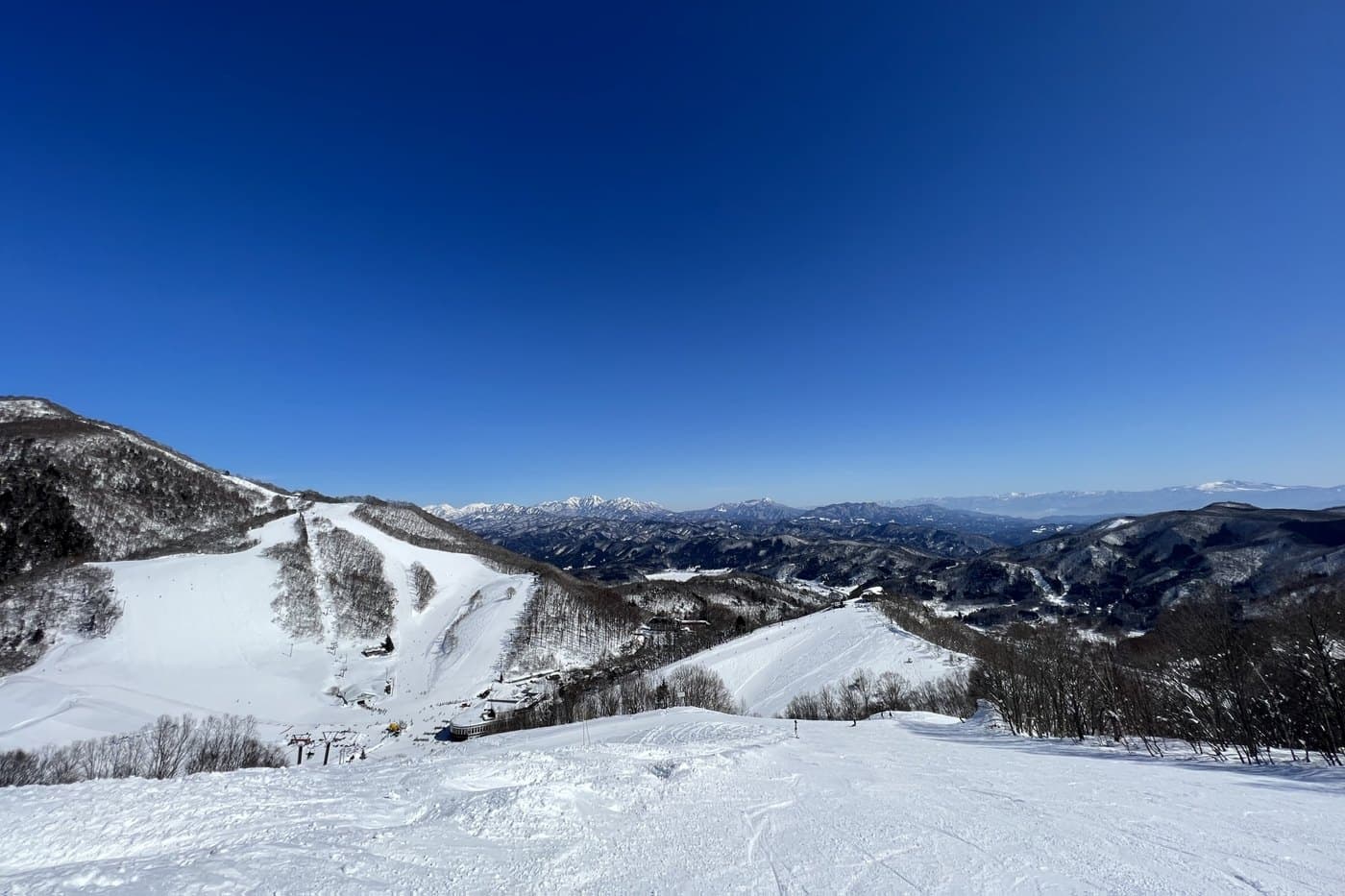 A wide groomed slope in Hakuba Valley on a clear day, with the snow-covered peaks of the Northern Japan Alps on the horizon