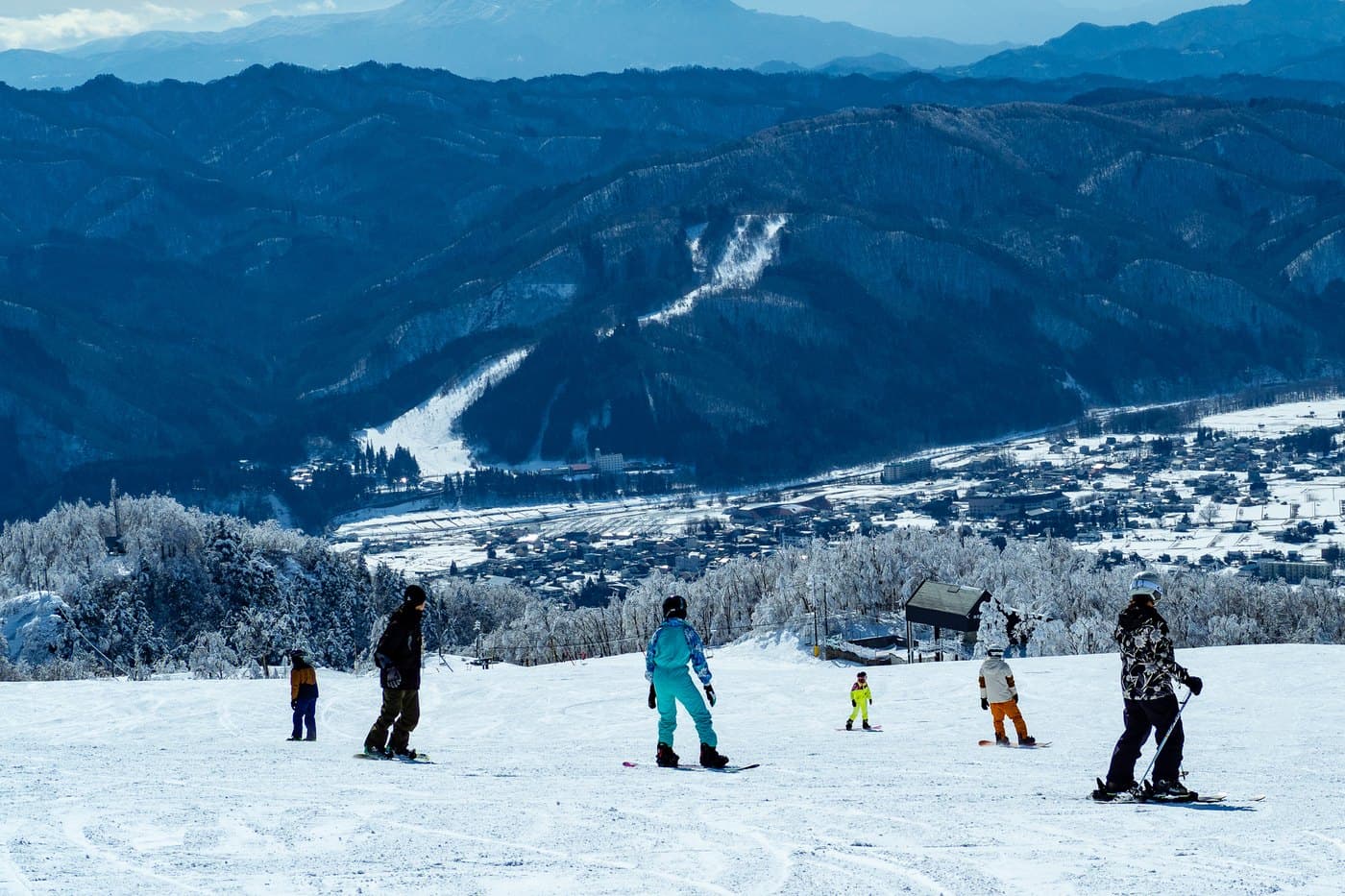 Skiers and snowboarders on a groomed run above Hakuba village, with the snow-covered valley floor and surrounding mountains below