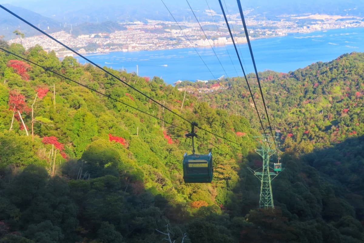 A Miyajima Ropeway gondola descending over forested slopes with autumn maples, the sea and the mainland coast below