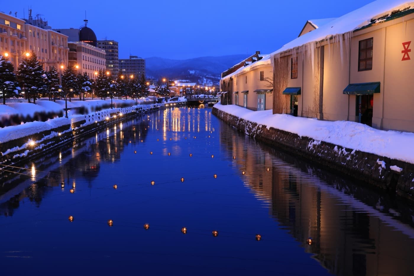 The Otaru Canal at blue hour in winter, candle lanterns floating on the water and snow piled on the old stone warehouses along the bank