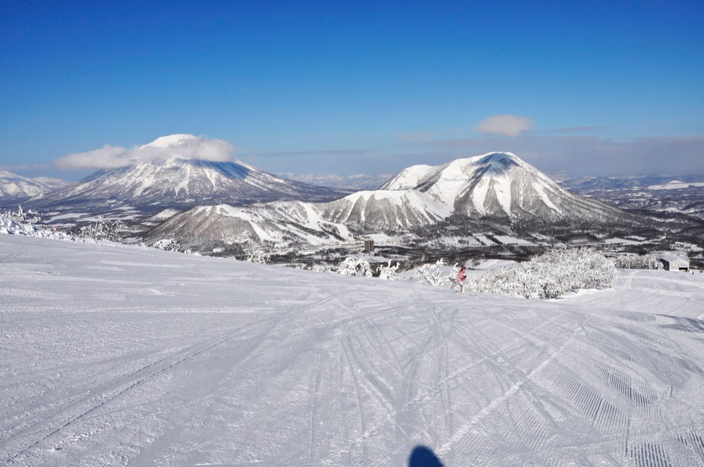 Groomed ski slope at Rusutsu Resort on a clear winter day, with a lone skier and the snow-capped cone of Mount Yotei on the horizon