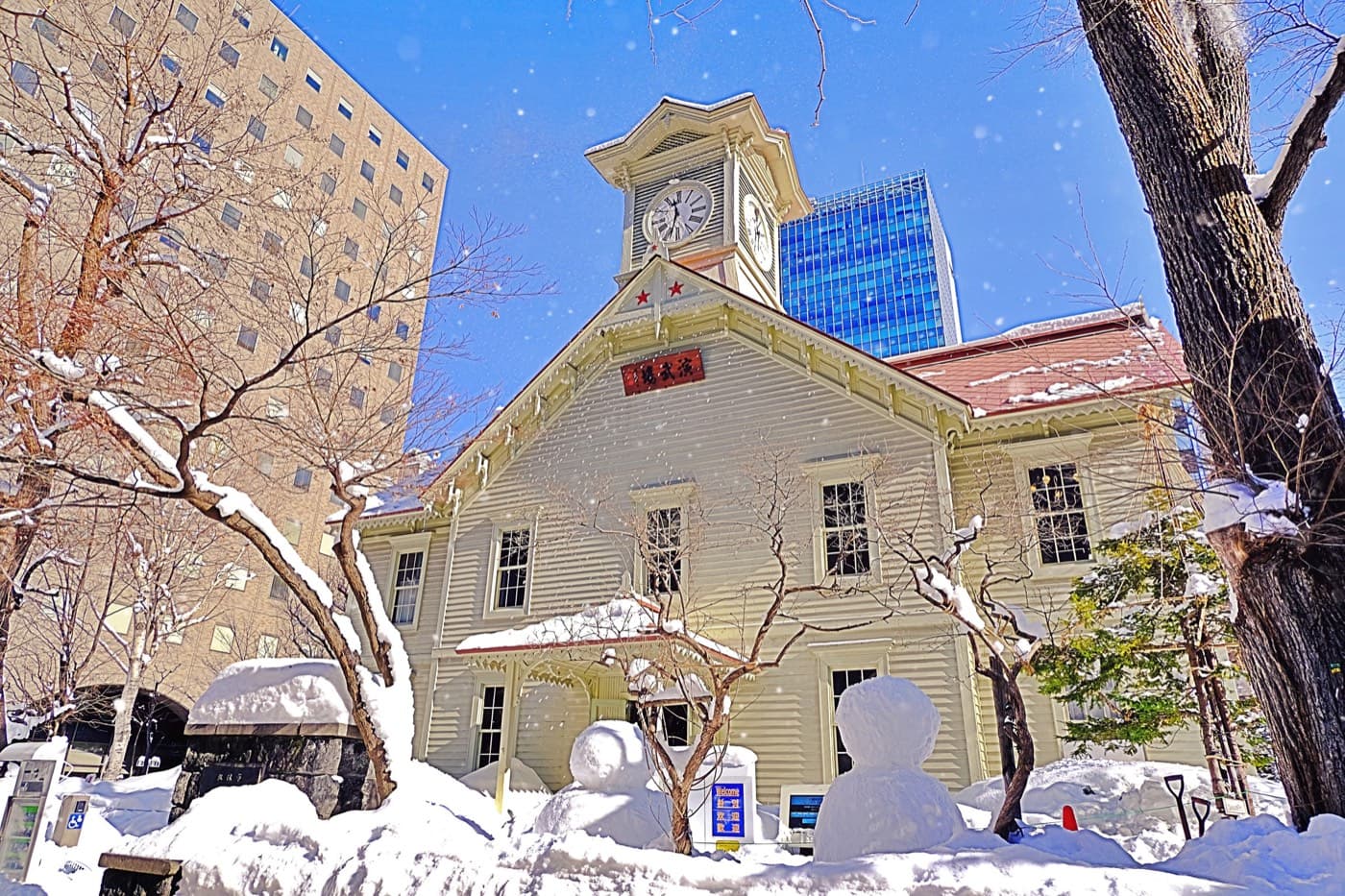 The historic wooden Sapporo Clock Tower under deep snow with a small snowman in front and modern towers behind