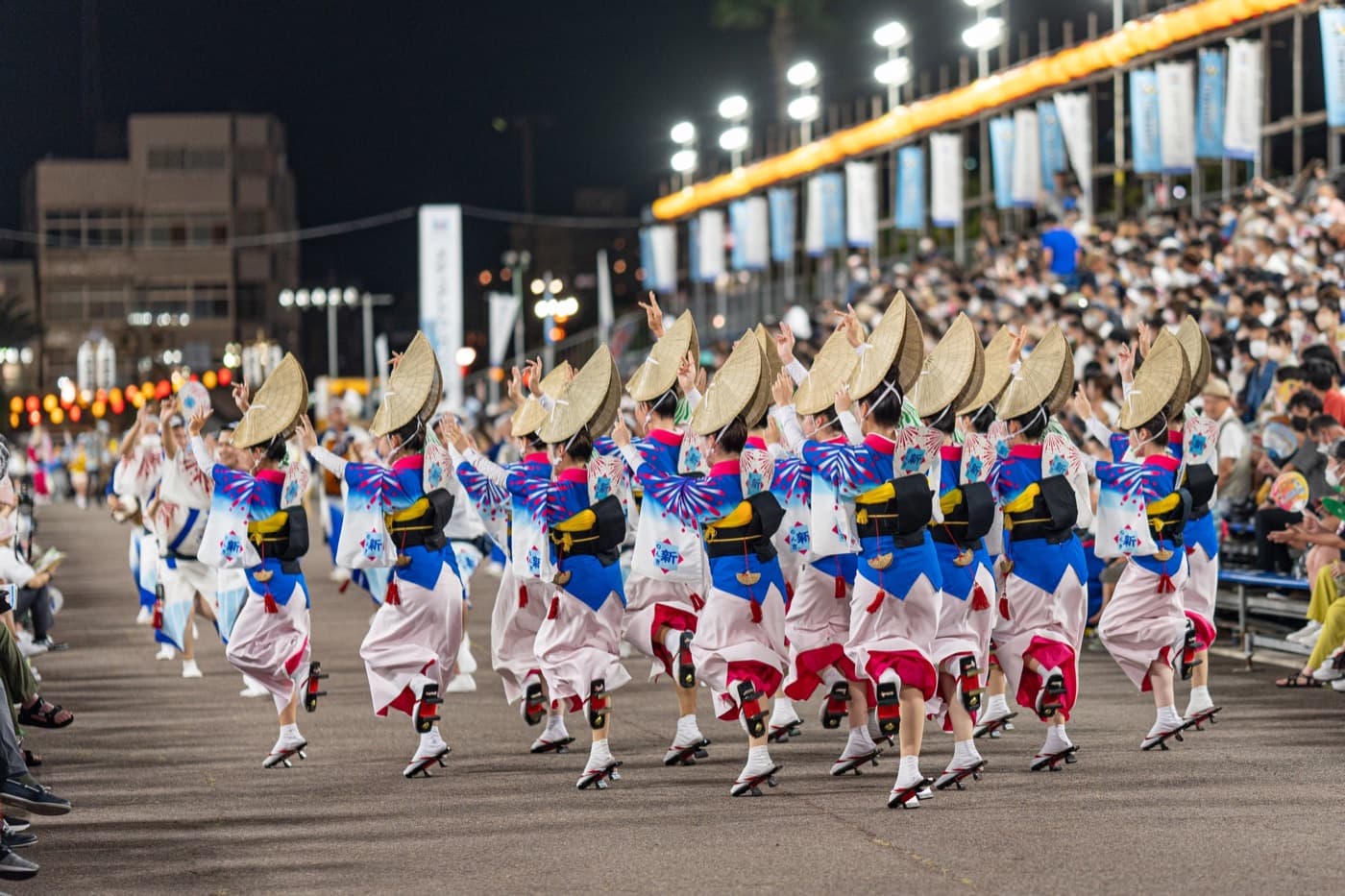 Awa Odori dancers in straw hats and blue-and-white yukata performing before a packed grandstand at night