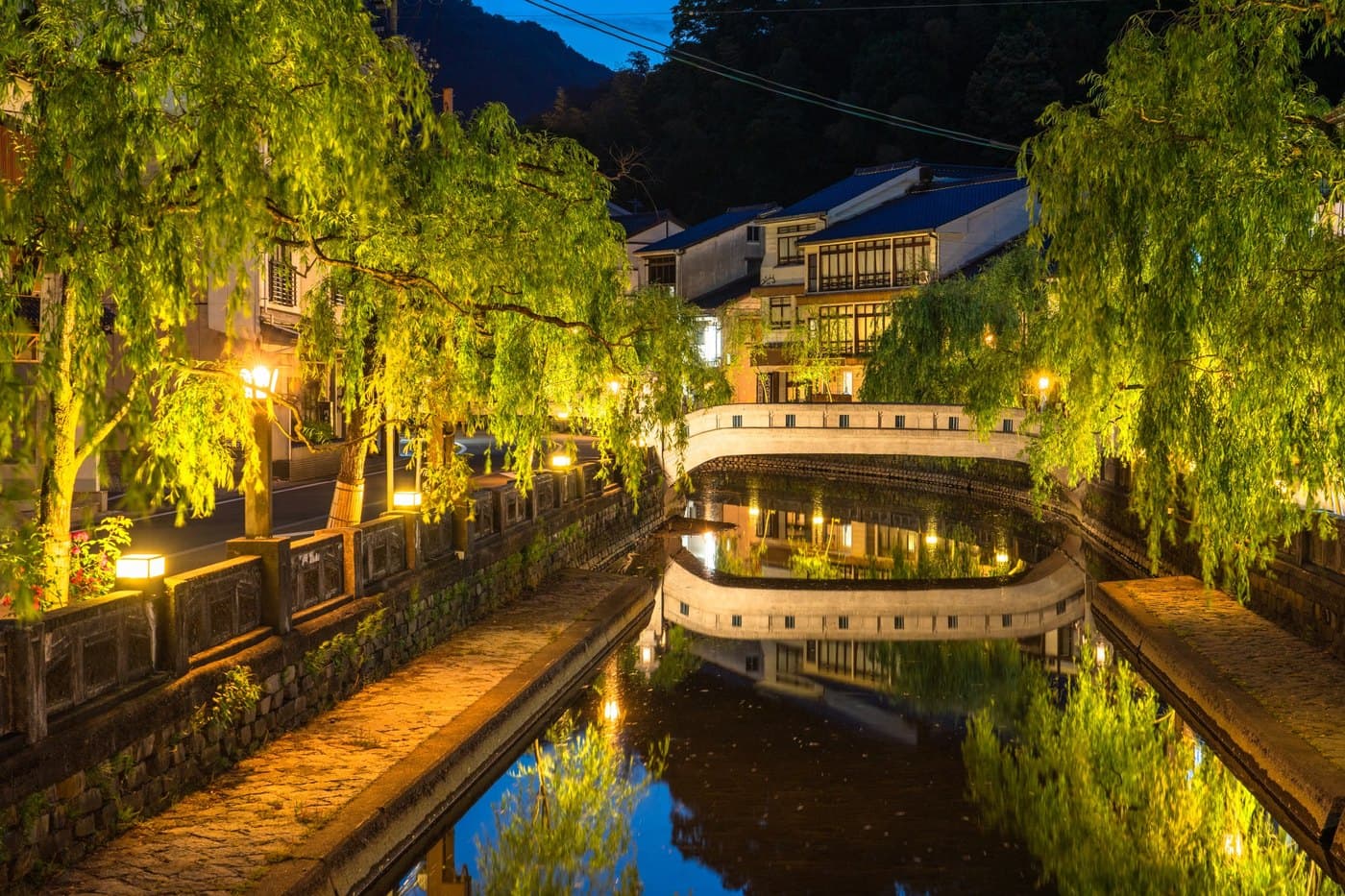 Kinosaki Onsen canal at night, lamps glowing along the willow-lined banks and the arched bridge reflected in the still water
