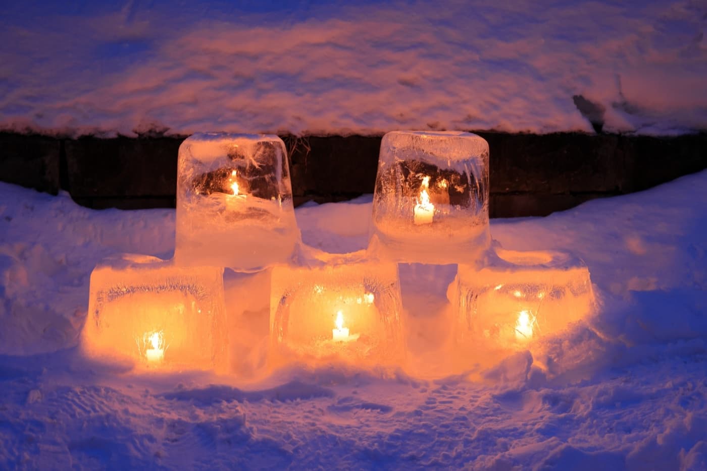 Five hand-made ice lanterns in the snow at dusk, each holding a lit candle, glowing orange against blue snow