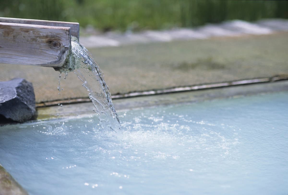 Milky white hot spring water pouring from a wooden spout into an open-air bath