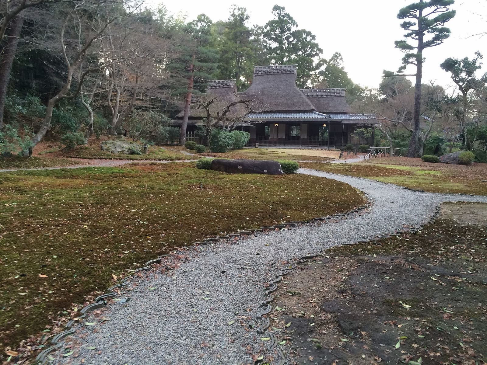A gravel path curving through the moss garden at Yoshikien toward a thatched-roof tea house in Nara
