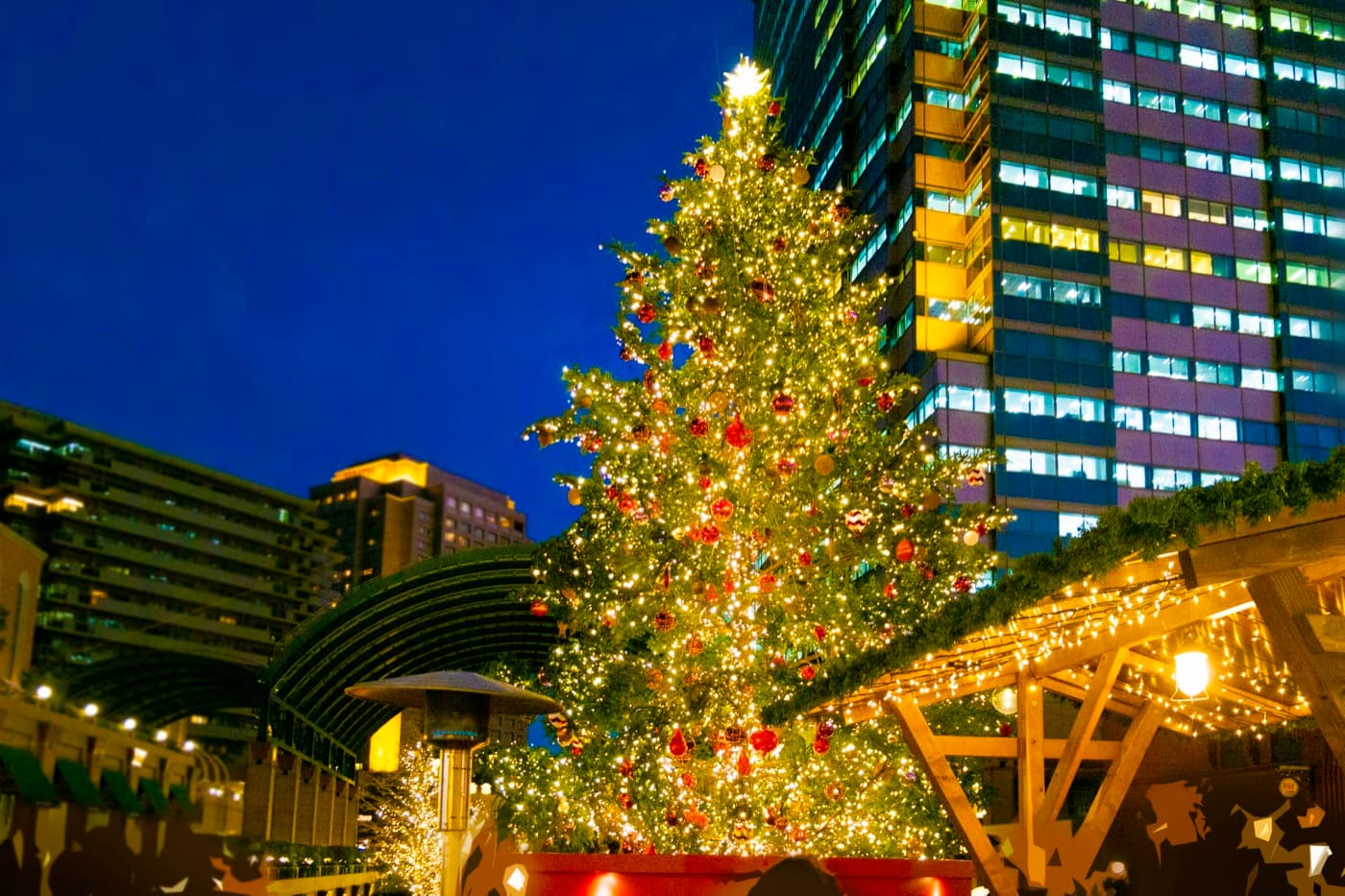 A tall decorated Christmas tree lit against a deep blue evening sky, with fairy-lit wooden market huts and a patio heater in the foreground