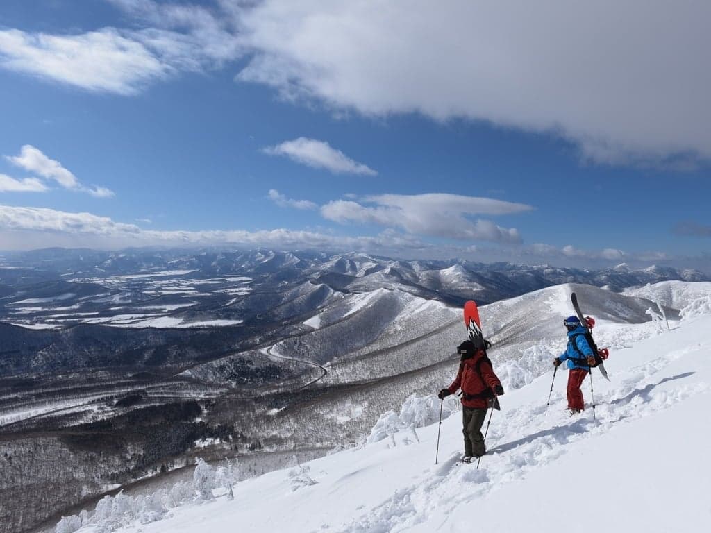 Two people hiking up a snowy ridge with skis on their packs, Hokkaido mountains stretching to the horizon