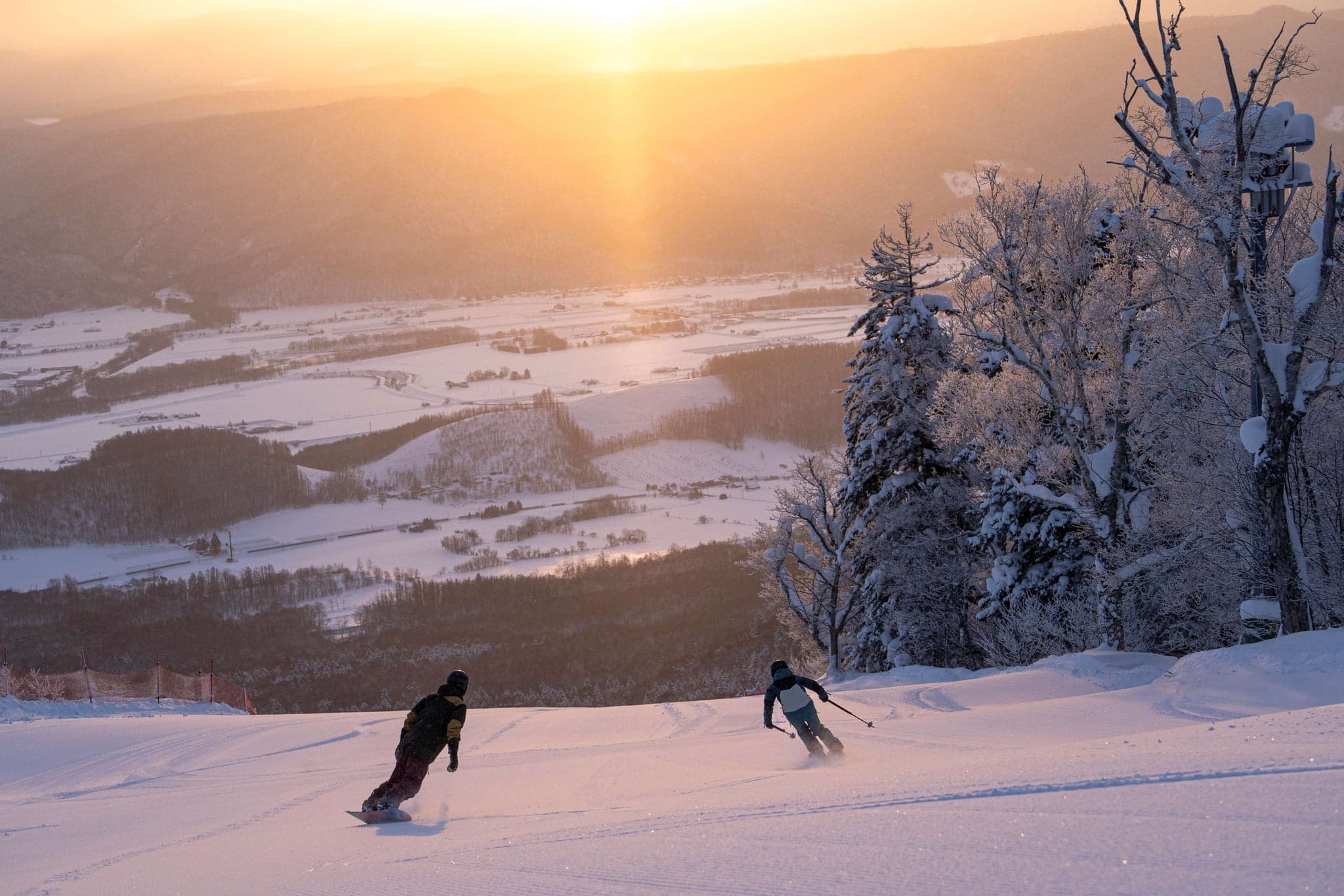 Two snowboarders riding a groomed Furano slope at sunset, with the snowy Furano basin and forest below
