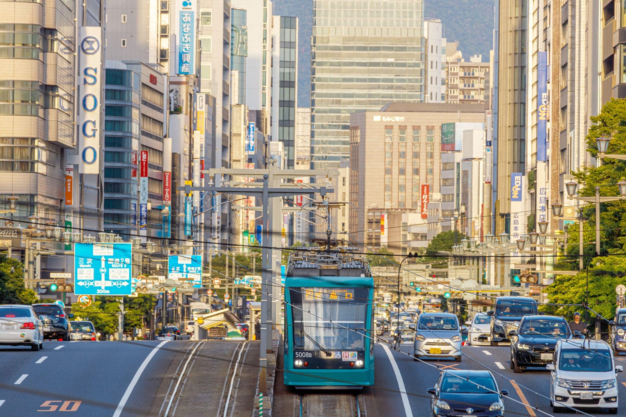 A green Hiroden streetcar running down a central Hiroshima avenue lined with department stores and office buildings
