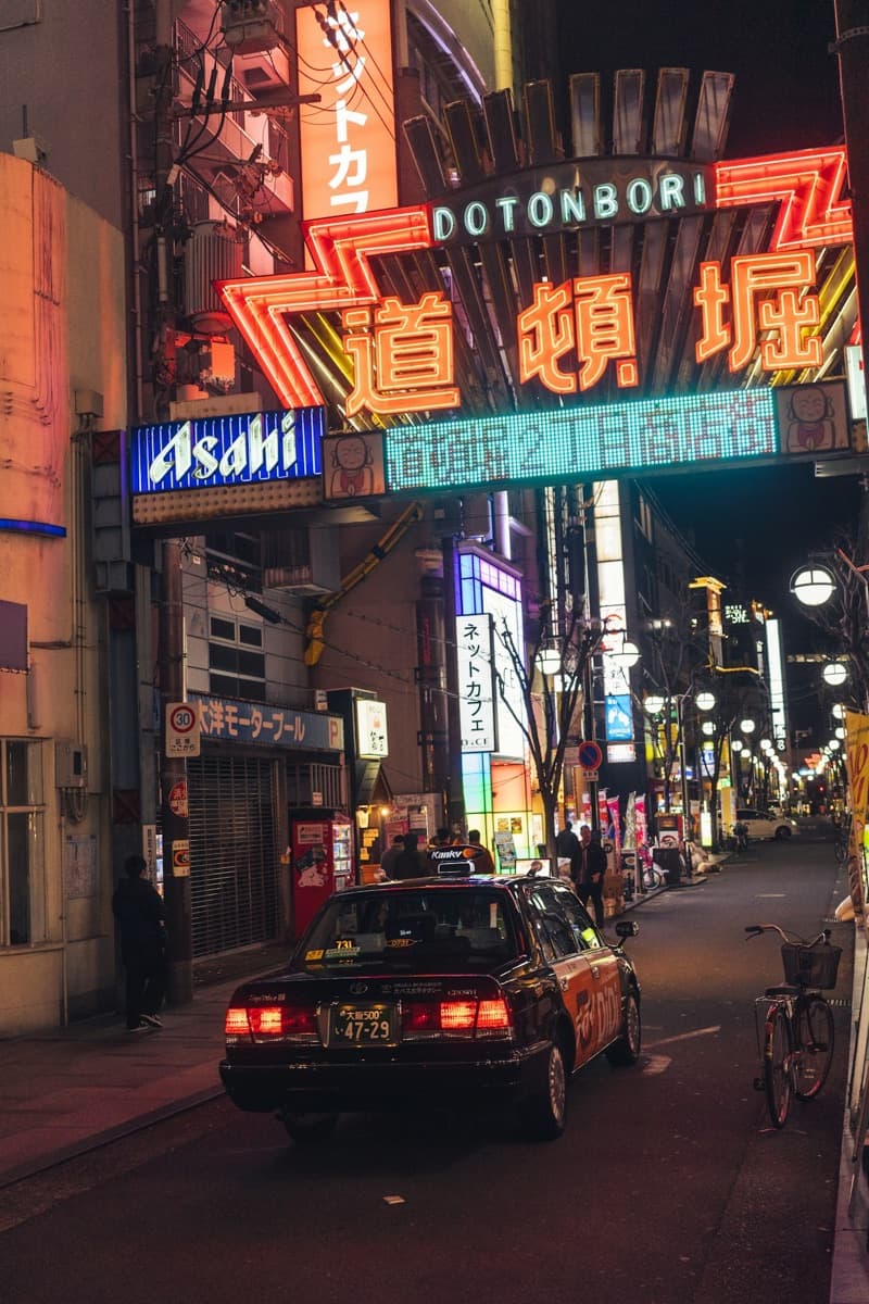 The Dotonbori entrance sign lit in red and neon at night with a taxi on the street below