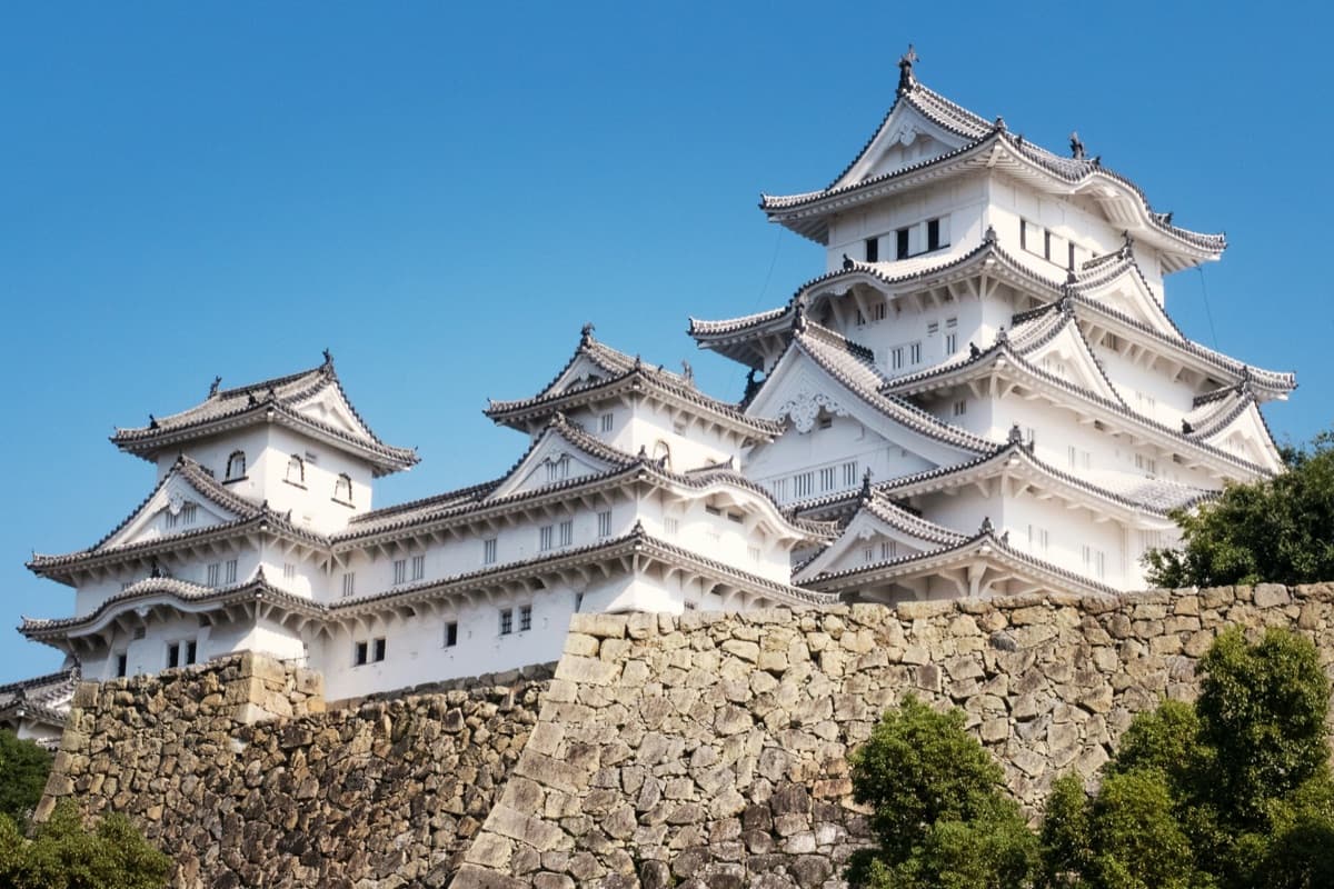 The white multi-tiered castle keep at Himeji rising above high stone walls and green trees under a blue sky