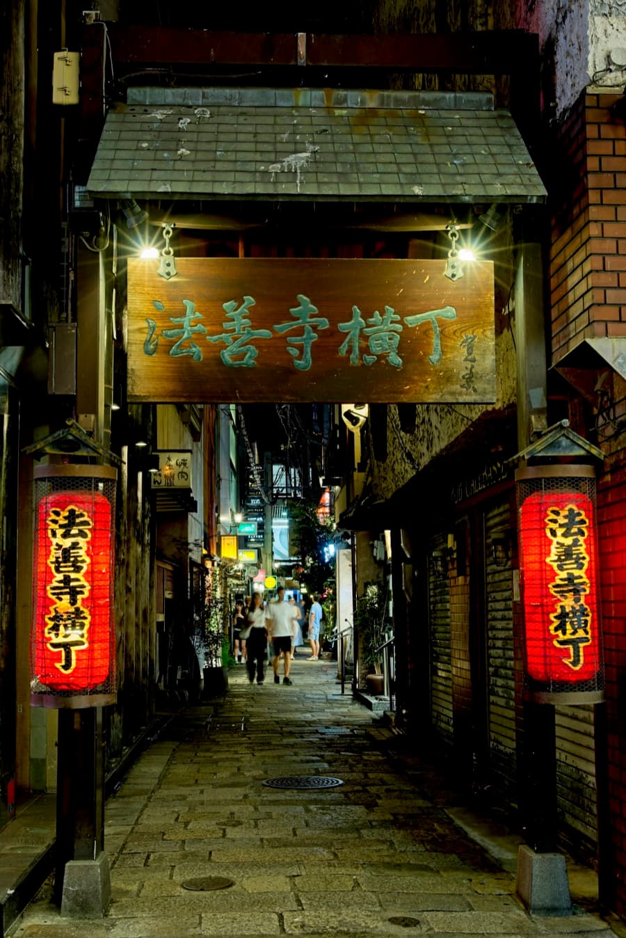 The narrow stone-paved Hozenji Yokocho alley at night with red lanterns and a wooden sign over the entrance