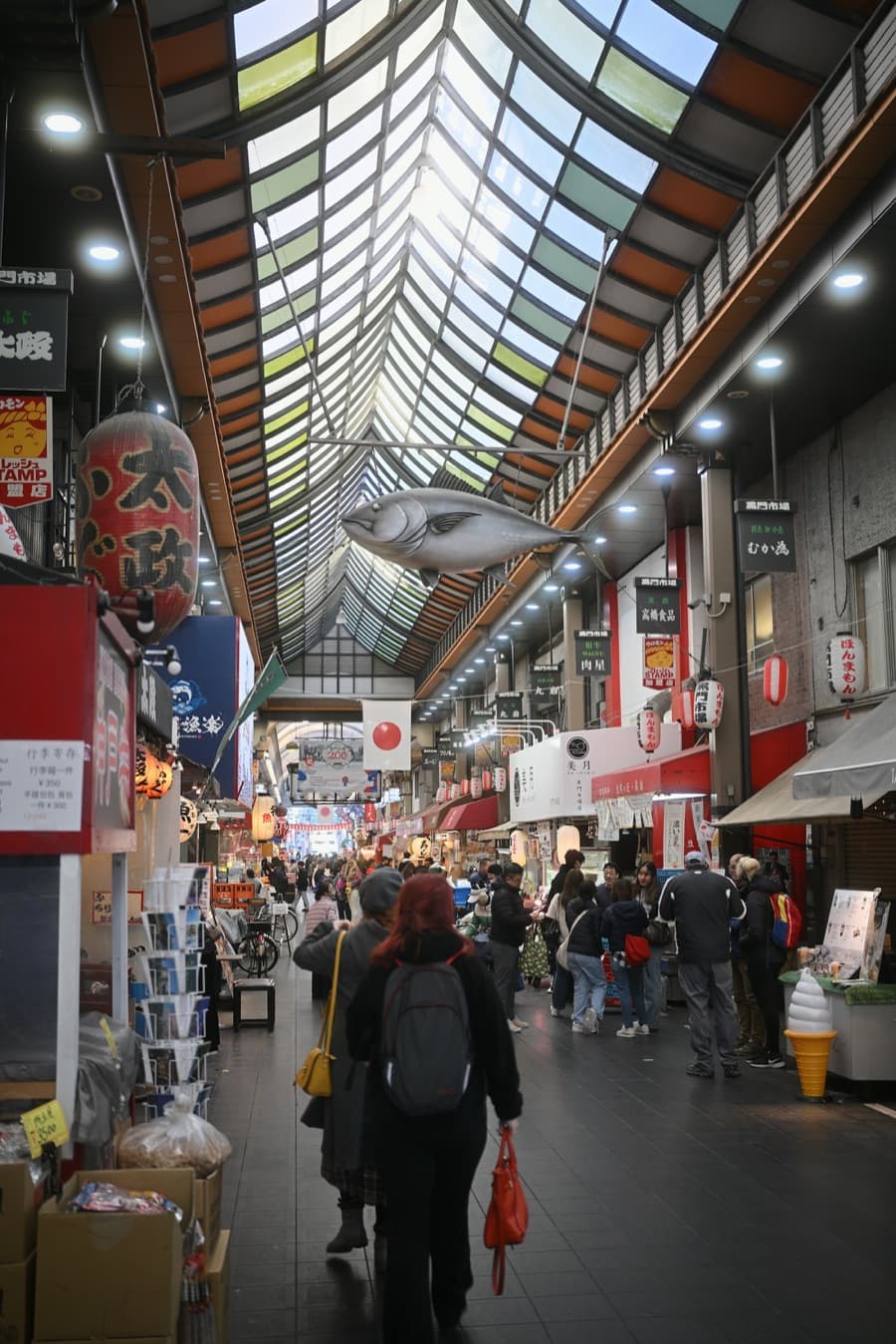Shoppers walking under the glass roof of Kuromon Market past shops, with a model fish hanging overhead