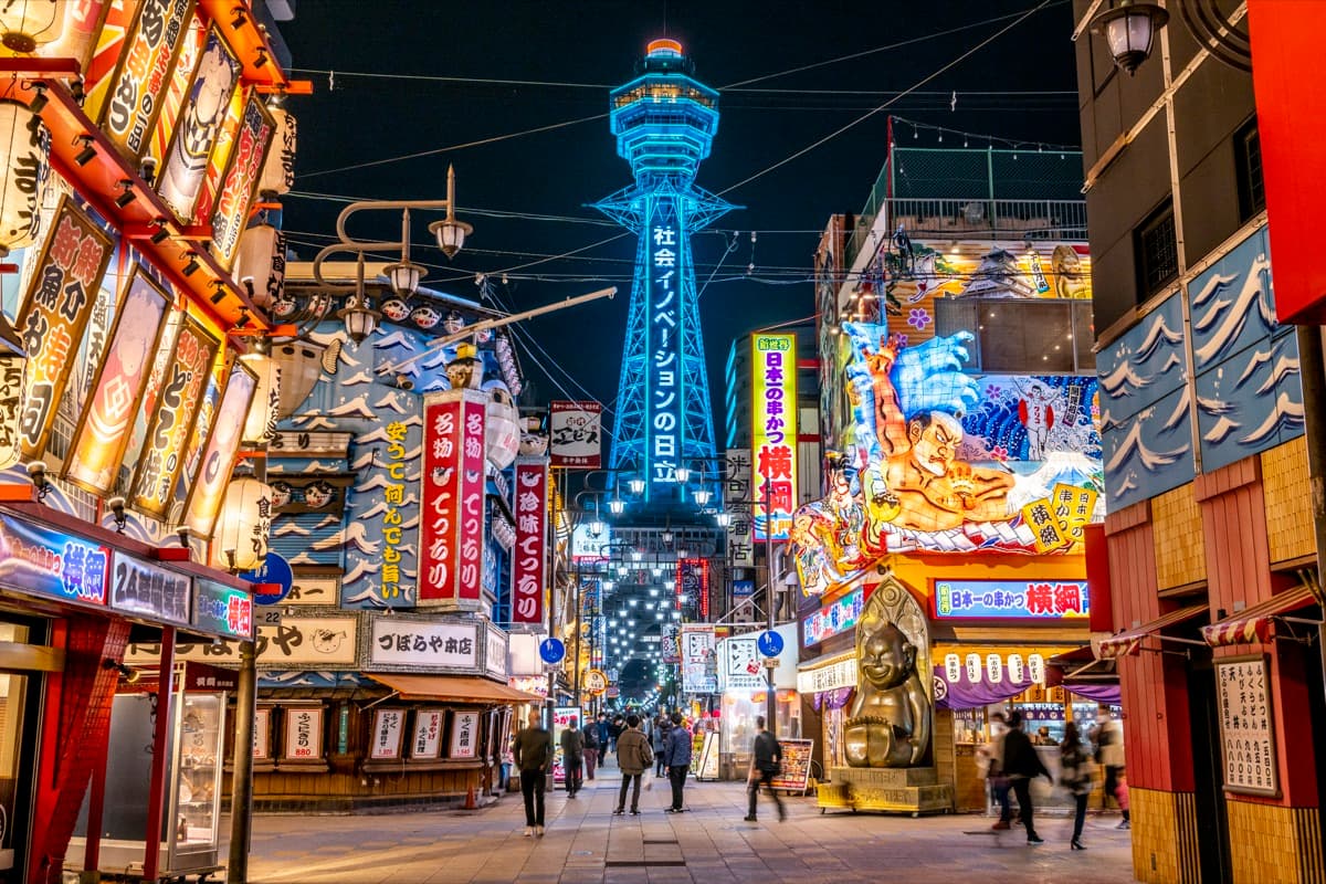 Tsutenkaku tower lit up in blue at night above a Shinsekai street lined with neon signs and restaurant fronts