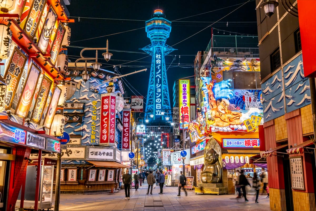 Shinsekai at night with Tsutenkaku tower lit in blue above a street of neon restaurant signs